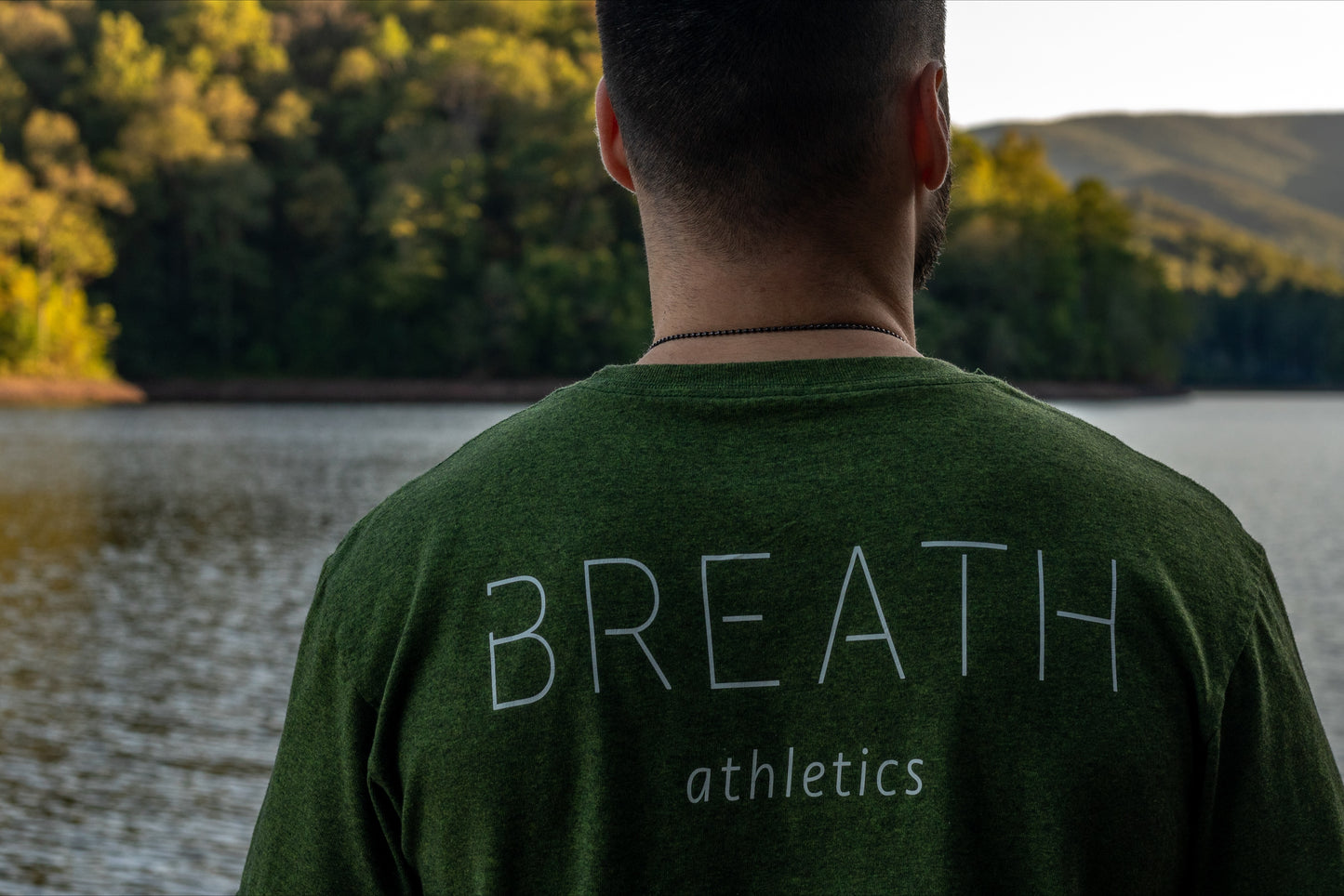 A person in a green t shirt facing a lake with the upper back of their green t shirt showing the brand name "Breath".