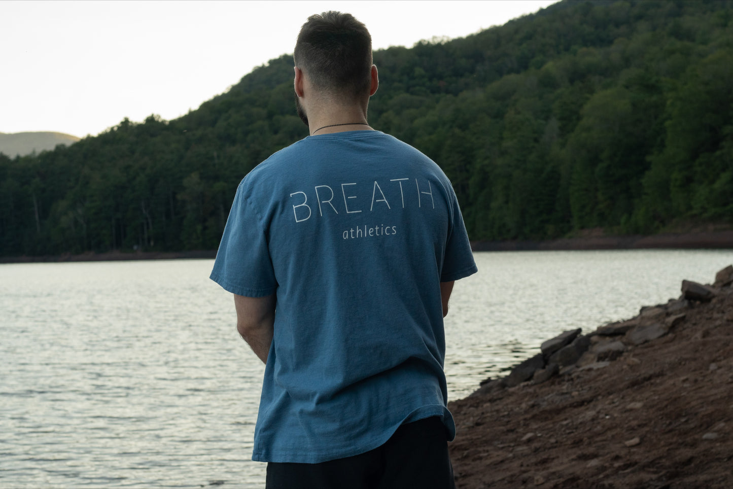 A person looking off into a beautiful lake wearing the light blue graphic t-shirt made with 100% organic cotton from breath showing the brand name across the back.