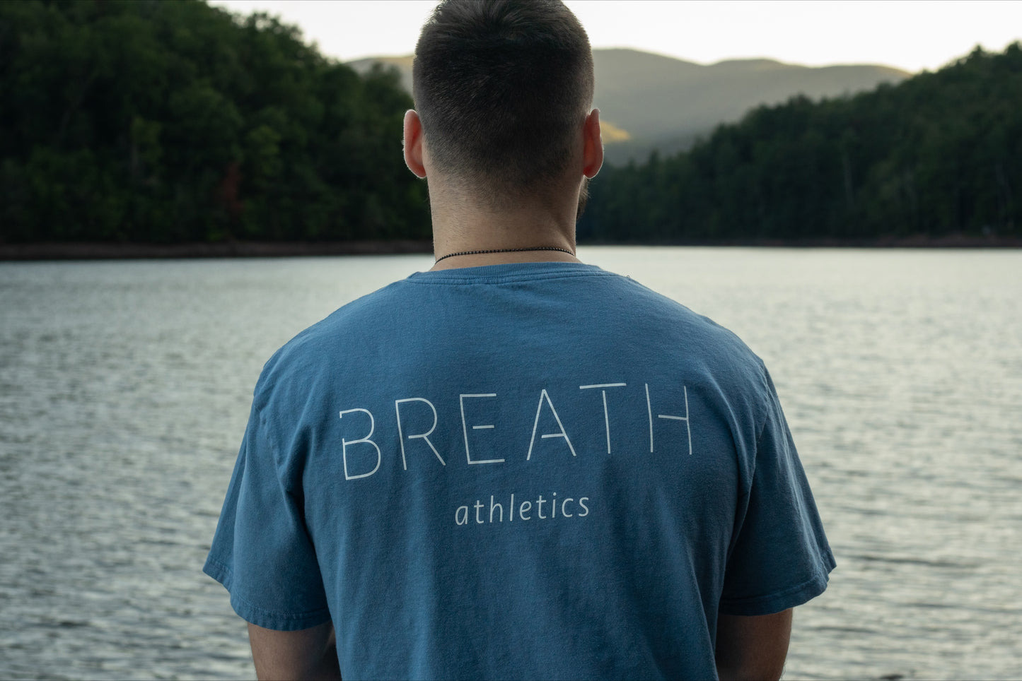 A person looking off into a beautiful lake wearing the light blue graphic t-shirt made with 100% organic cotton from breath showing the brand name across the back.