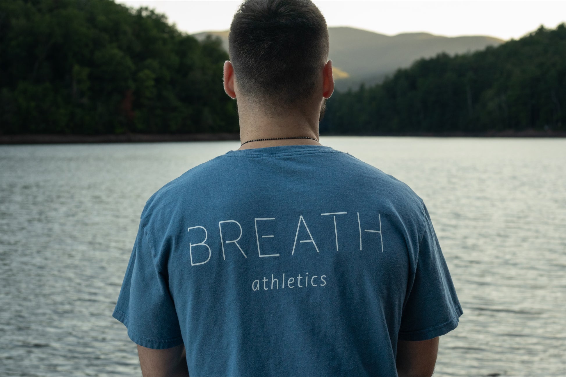 A person looking off into a beautiful lake wearing the light blue graphic t-shirt made with 100% organic cotton from breath showing the brand name across the back.