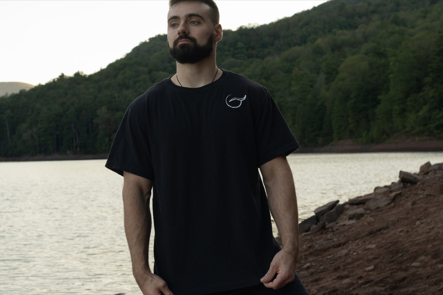 A person displaying the front of the 100% Organic cotton black graphic t shirt by Breath with the logo on the left chest and the brand name across the back, standing on the shore of a beautiful lake.