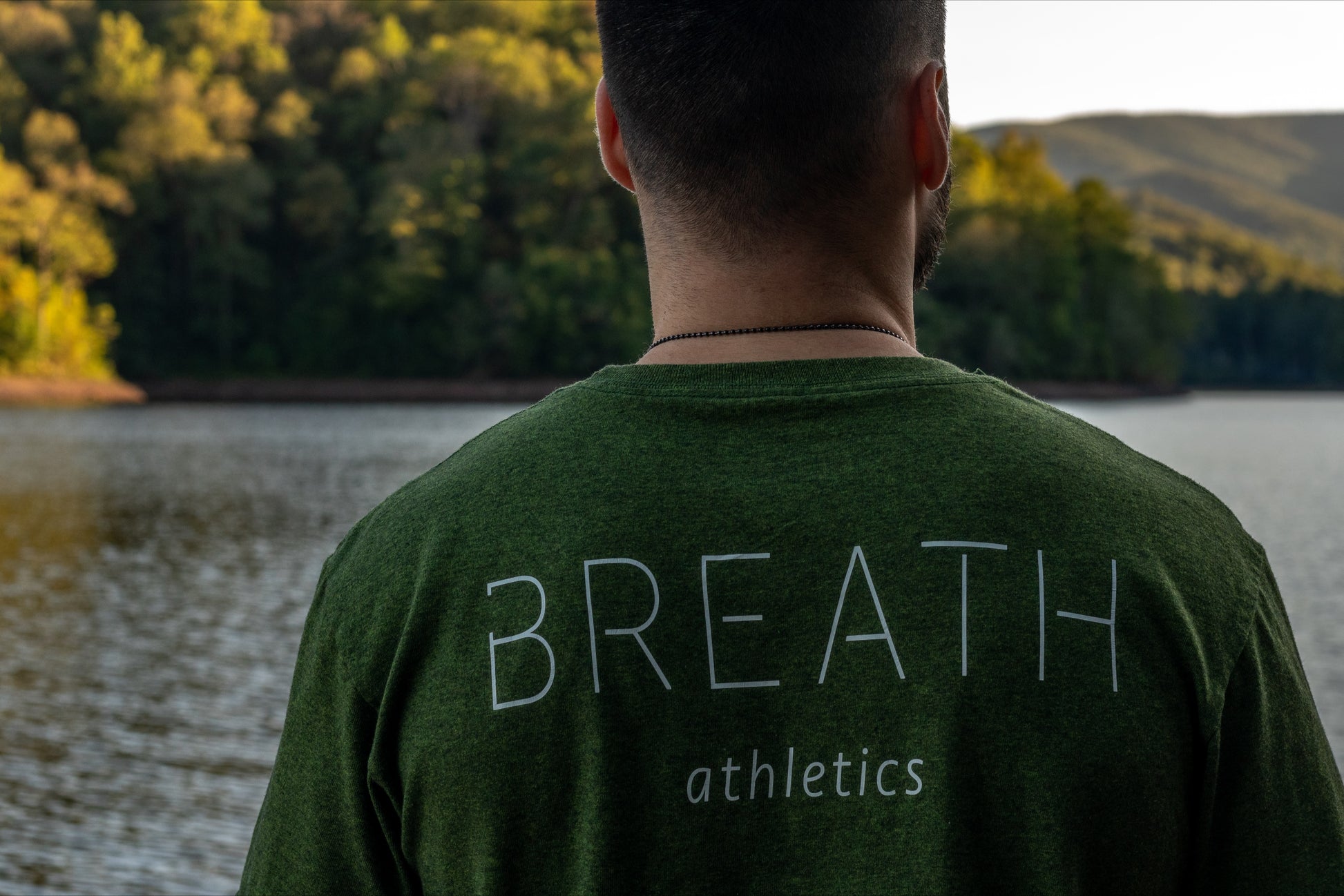 A person in a green t shirt facing a lake with the upper back of their green t shirt showing the brand name "Breath".