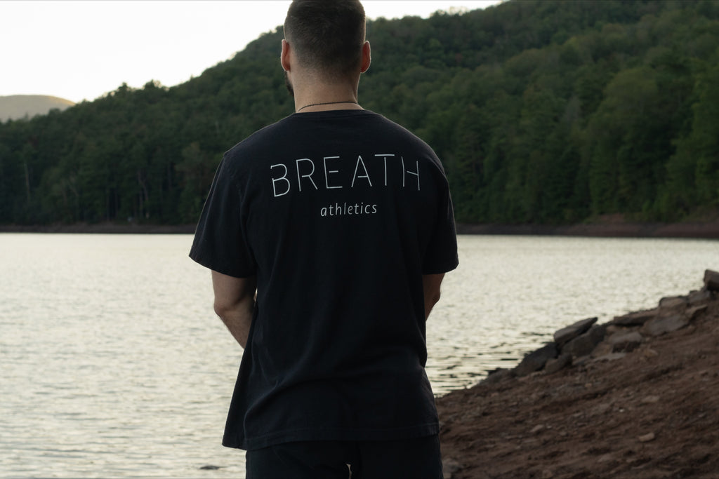 A person displaying the back of the 100% Organic cotton black graphic t shirt by Breath with the logo on the left chest and the brand name across the back, standing on the shore of a beautiful lake.
